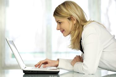 Woman in white jacket leaning on her desk looking at her laptop.